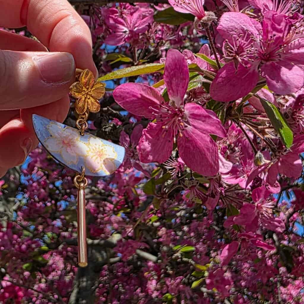 Boucle d'oreille demi-lune en papier japonais bleu avec motif sakura doré, tenue devant un cerisier en fleurs fuchsia