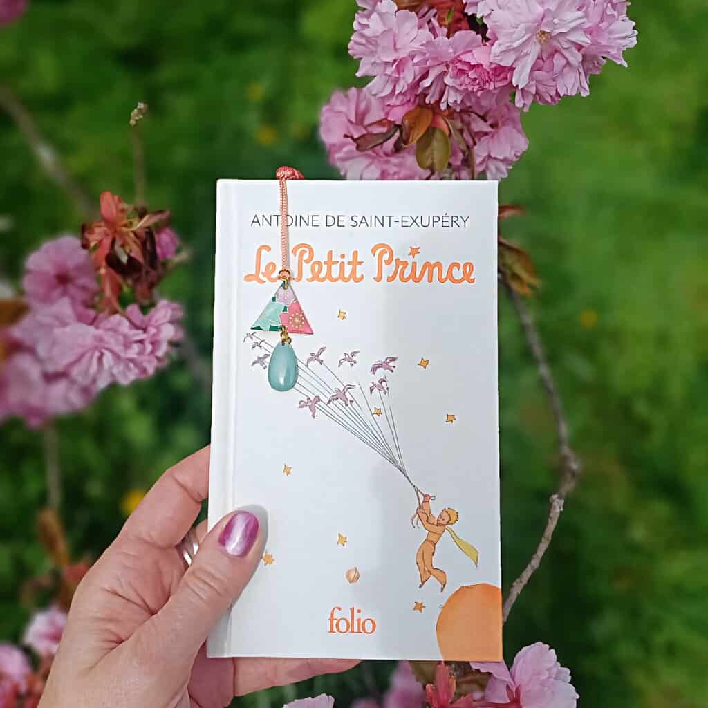 Livre Le Petit Prince tenu à la main avec un marque-pages triangle en papier japonais fleuri, devant un fond de cerisiers roses en fleurs